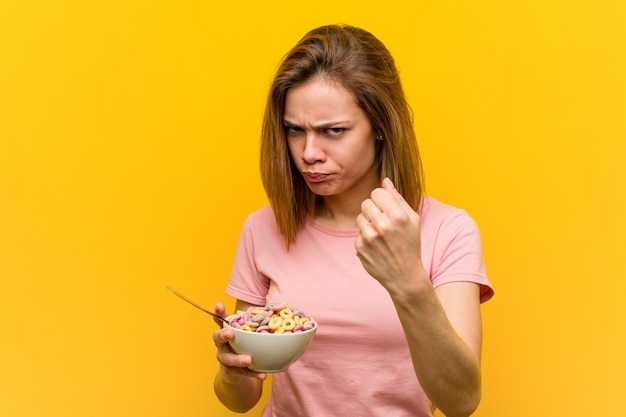 Giovane donna che tiene una ciotola di cereali che mostra pugno alla macchina fotografica, espressione facciale aggressiva.