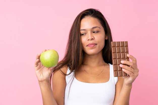 Giovane donna che tiene una mela e un cioccolato sopra la parete isolata