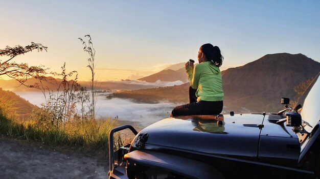 Giovane donna che tiene una tazza di bevanda calda e si siede sul cofano dell'auto mentre si gode la vista sulle montagne di Batur all'ora dell'alba