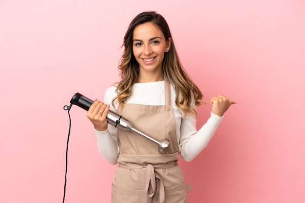 Giovane donna che usa il frullatore a immersione su sfondo rosa isolato che punta al lato per presentare un prodotto