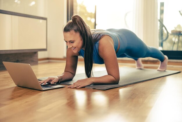 Giovane donna che usa il laptop mentre fa esercizi di plancia a casa al mattino.