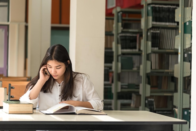 Giovane donna che usa il telefono mentre è seduta accanto al libro sulla scrivania della biblioteca