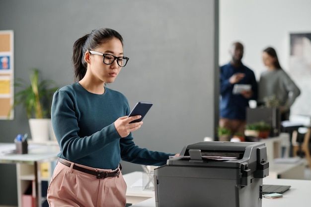 Giovane donna che usa lo smartphone mentre stampa documenti