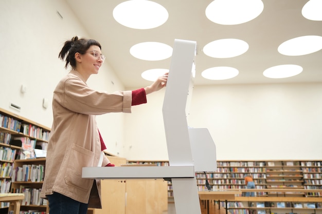 Giovane donna che utilizza attrezzature speciali e sceglie un libro in biblioteca