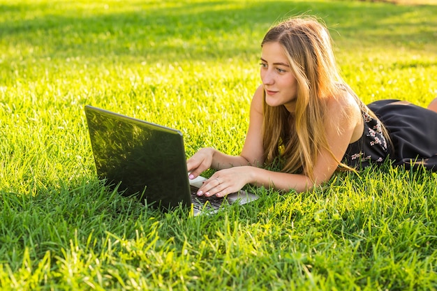 Giovane donna che utilizza computer portatile nel parco sdraiato sull'erba verde. Concetto di attività per il tempo libero.