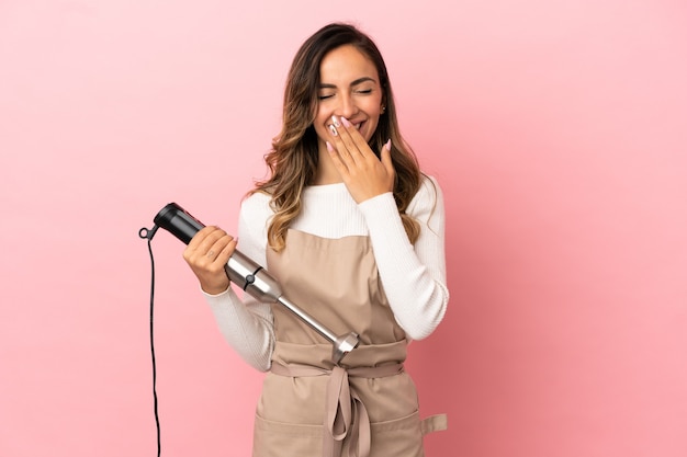 Giovane donna che utilizza frullatore a immersione su sfondo rosa isolato felice e sorridente che copre la bocca con la mano