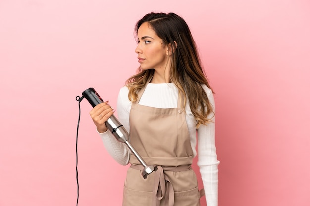 Giovane donna che utilizza frullatore a immersione su sfondo rosa isolato guardando al lato