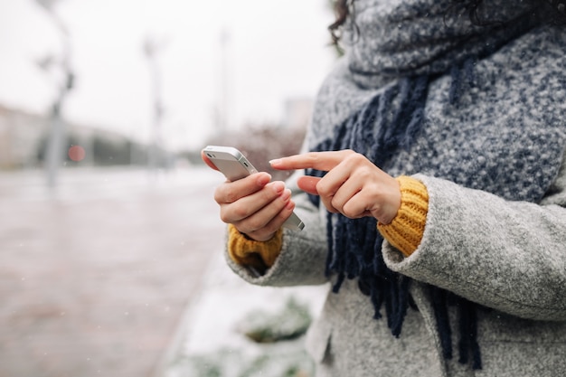 Giovane donna che utilizza il suo telefono cellulare in un parco invernale innevato. Primo piano della femmina che controlla le notizie e che manda un sms sul suo cellulare all'aperto durante la fredda stagione invernale. Concetto di gadget delle persone.