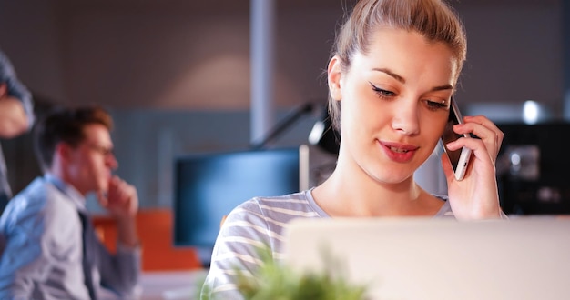 Giovane donna che utilizza il telefono cellulare mentre si lavora al computer di notte in un ufficio buio.