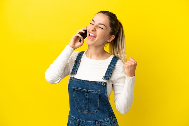 Giovane donna che utilizza il telefono cellulare su sfondo giallo isolato che celebra una vittoria