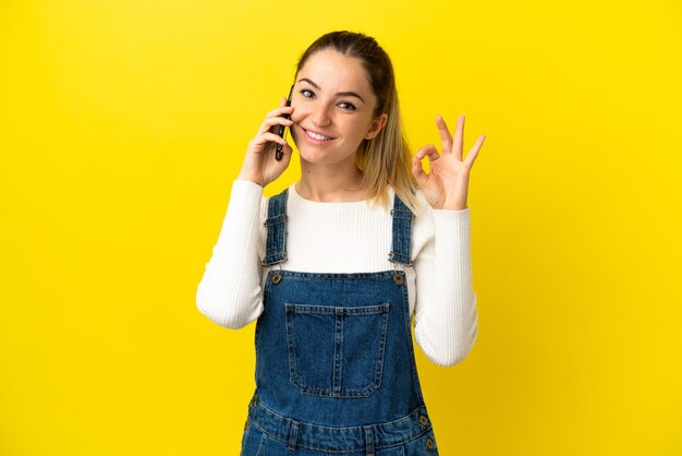 Giovane donna che utilizza il telefono cellulare su sfondo giallo isolato che mostra segno ok con le dita
