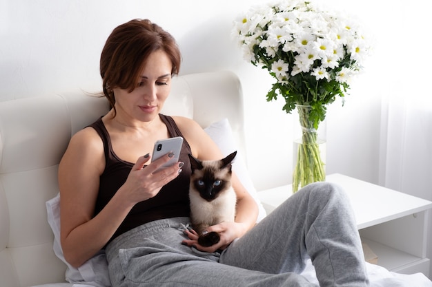 Giovane donna che utilizza il telefono con il suo gatto siamese in camera da letto con fiori.