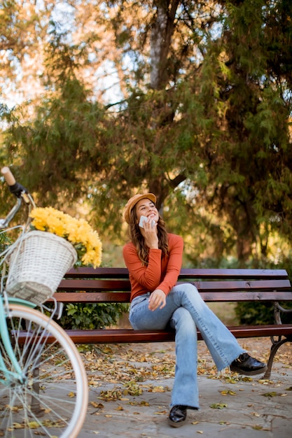 Giovane donna che utilizza telefono nel parco di autunno che si siede sul banco in bicicletta
