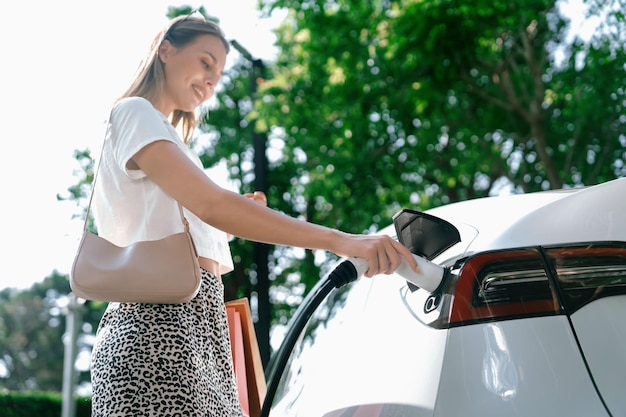 Giovane donna che viaggia con un'auto elettrica EV in carica nel giardino all'aperto di una città verde e sostenibile in estate Stile di vita urbano sostenibile grazie all'energia verde pulita e ricaricabile delle interiora dei veicoli elettrici BEV