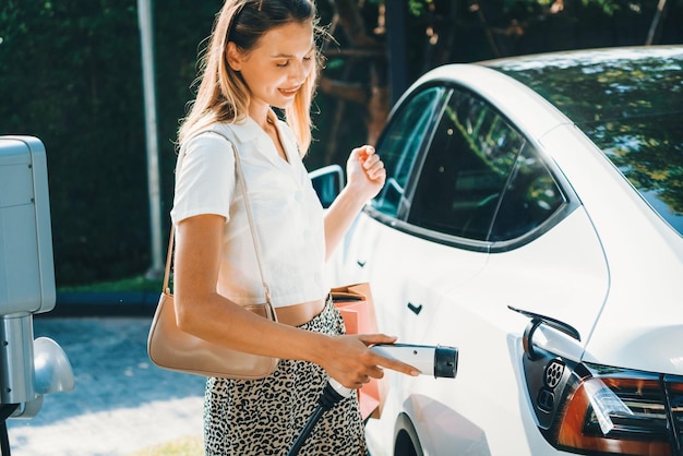 Giovane donna che viaggia con un'auto elettrica EV in carica nel giardino all'aperto di una città verde e sostenibile in estate Stile di vita urbano sostenibile grazie all'energia verde pulita e ricaricabile delle interiora dei veicoli elettrici BEV