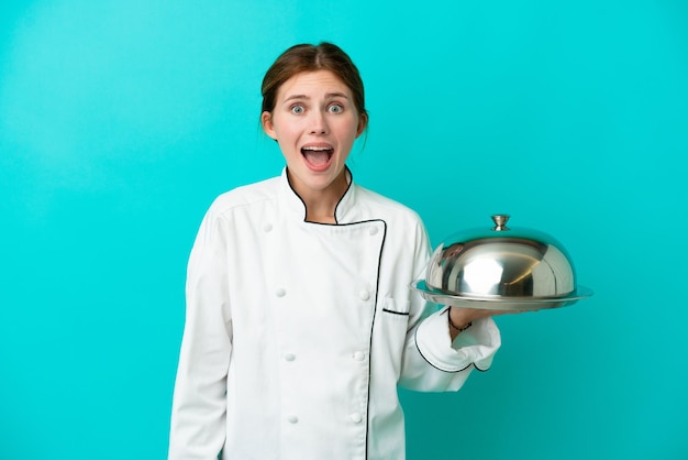Giovane donna chef con vassoio isolato su sfondo blu con espressione facciale a sorpresa