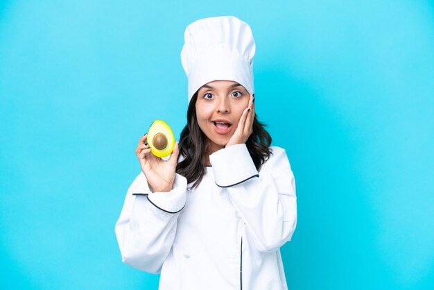 Giovane donna chef ispanica che tiene avocado isolato su sfondo blu con espressione facciale sorpresa e scioccata