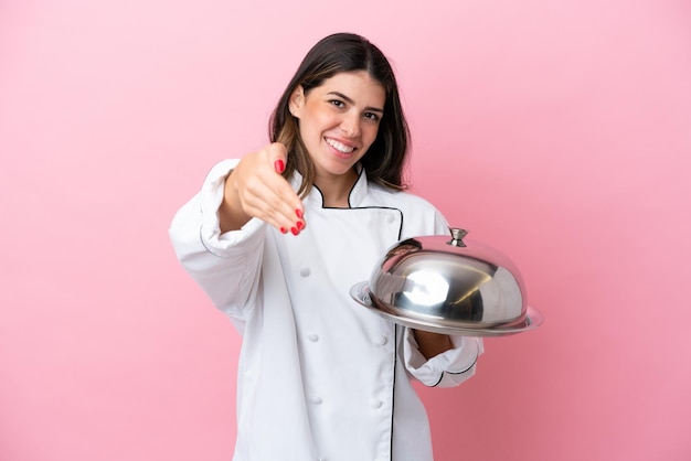 Giovane donna chef italiana che tiene vassoio con coperchio isolato su sfondo rosa che stringe la mano per chiudere un buon affare