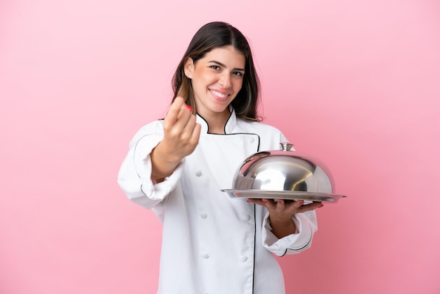 Giovane donna chef italiana che tiene vassoio con coperchio isolato su sfondo rosa facendo un gesto in arrivo