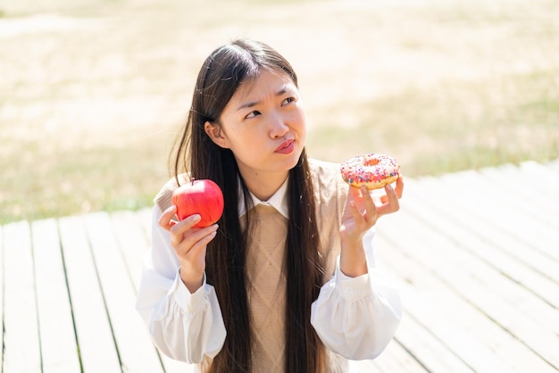 Giovane donna cinese all'aperto che tiene mela e ciambella e ha dubbi