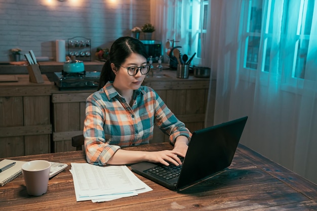 giovane donna cinese asiatica elegante lavoratore freelance che lavora al computer portatile a casa di notte al buio. bella signora che scrive sulla tastiera del pc del taccuino con il documento sul tavolo in cucina a mezzanotte.