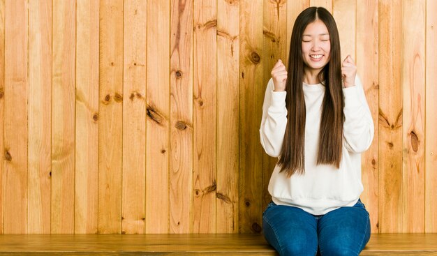 Giovane donna cinese che si siede su un posto di legno che alza pugno, sentendosi felice e riuscito. Concetto di vittoria.