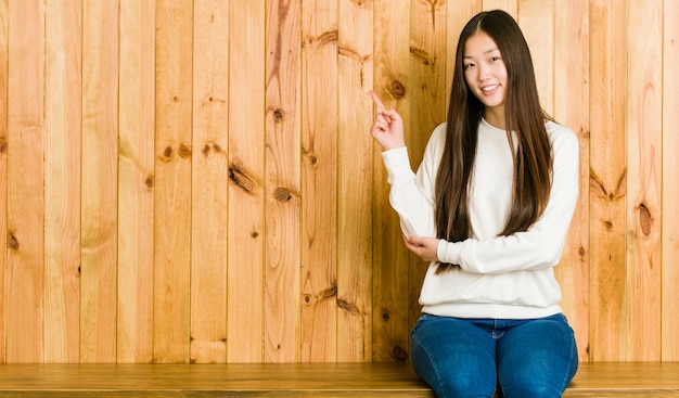 Giovane donna cinese che si siede su un posto di legno che sorride allegramente indicando con l'indice via.