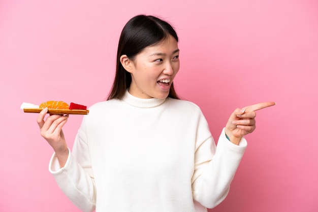 Giovane donna cinese che tiene sashimi isolato su sfondo rosa che punta il dito di lato e presenta un prodotto