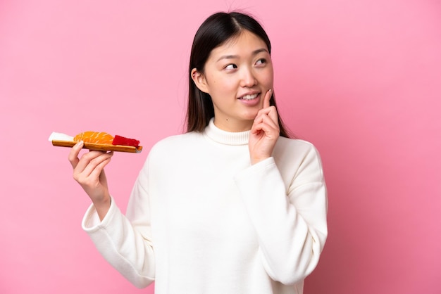 Giovane donna cinese che tiene sashimi isolato su sfondo rosa pensando a un'idea mentre guarda in alto