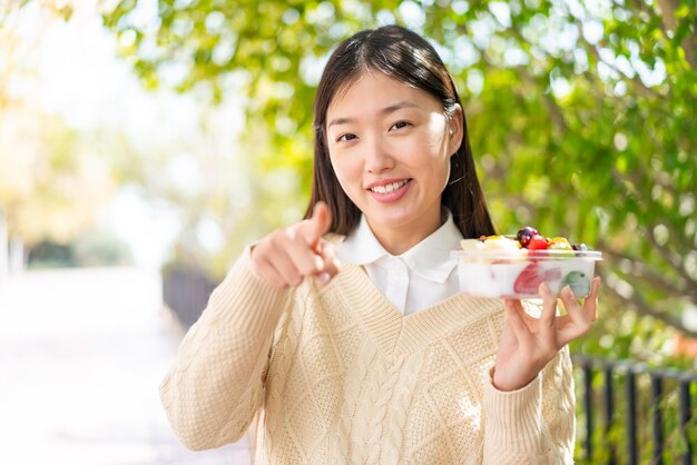 Giovane donna cinese che tiene una ciotola di frutta all'aperto sorpreso e che punta davanti