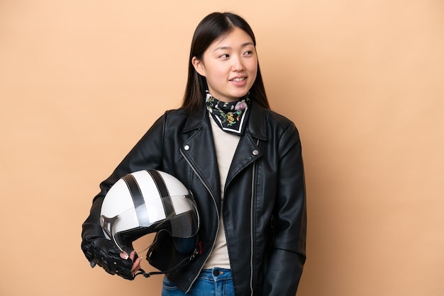 Giovane donna cinese con un casco da motociclista isolato su sfondo beige che guarda di lato e sorride