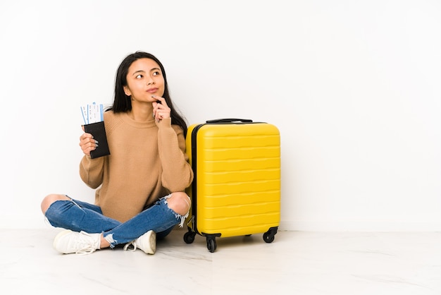 Giovane donna cinese del viaggiatore che si siede sul pavimento con una valigia isolata che osserva obliquamente con espressione dubbiosa e scettica.