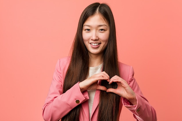 Giovane donna cinese di affari che porta vestito rosa che sorride e che mostra una figura del cuore con lui mani.