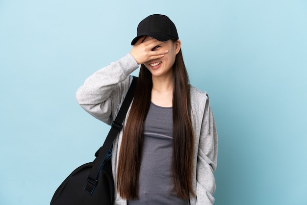 Giovane donna cinese di sport con la borsa di sport sopra gli occhi blu della copertura isolati dalle mani e dal sorridere