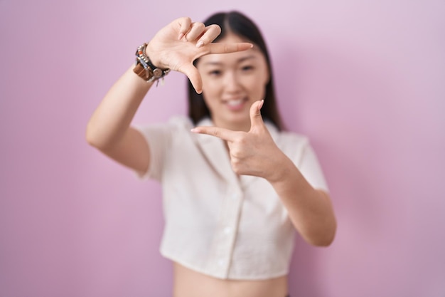 Giovane donna cinese in piedi su sfondo rosa sorridente che fa cornice con le mani e le dita con la creatività del viso felice e il concetto di fotografia