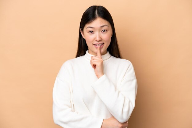 Giovane donna cinese isolata su sfondo beige che mostra un gesto di silenzio mettendo il dito in bocca