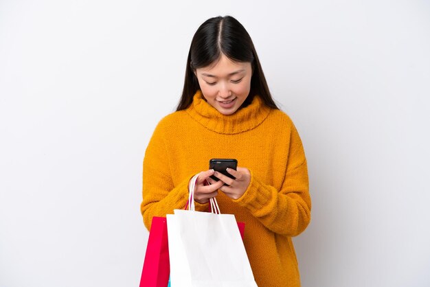 Giovane donna cinese isolata su sfondo bianco che tiene le borse della spesa e scrive un messaggio con il suo cellulare a un amico