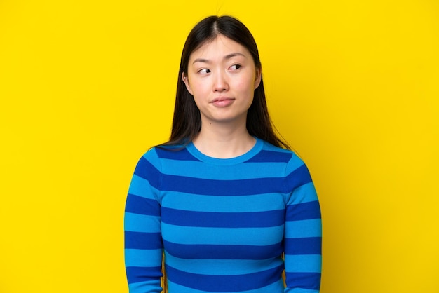 Giovane donna cinese isolata su sfondo giallo che fa un gesto di dubbi guardando il lato