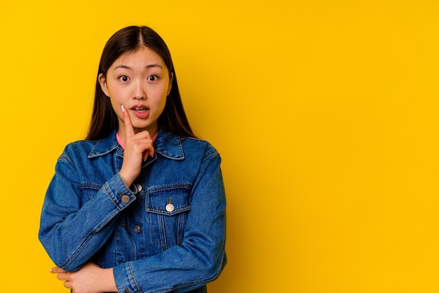 Giovane donna cinese isolata su sfondo giallo che guarda lateralmente con espressione dubbiosa e scettica.