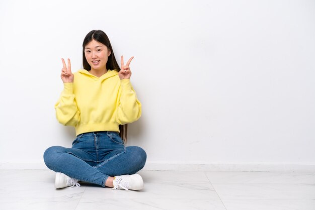 Giovane donna cinese seduta sul pavimento isolata sul muro bianco che mostra il segno della vittoria con entrambe le mani