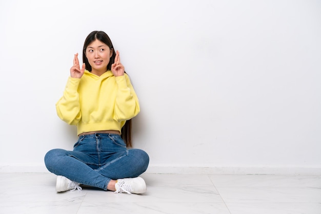 Giovane donna cinese seduta sul pavimento isolata sul muro bianco con le dita incrociate