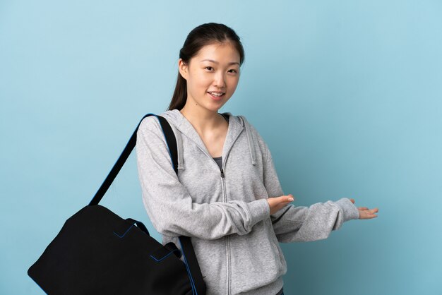 Giovane donna cinese sportiva con borsa sportiva sopra la parete blu isolata che estende le mani a lato per invitare a venire