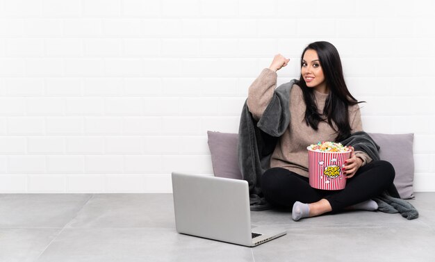 Giovane donna colombiana che tiene una ciotola di popcorn e che mostra un film in un computer portatile che fa gesto forte