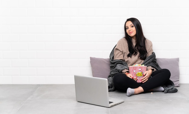 Giovane donna colombiana che tiene una ciotola di popcorn e che mostra un film in un computer portatile con le braccia incrociate e guardando avanti