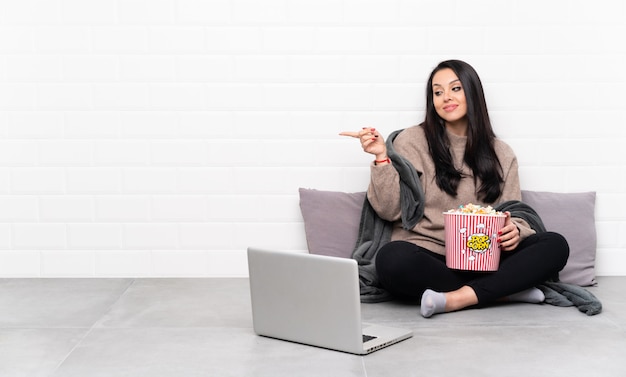 Giovane donna colombiana che tiene una ciotola di popcorn e che mostra un film in un computer portatile tenendo copyspace immaginario sul palmo da inserire