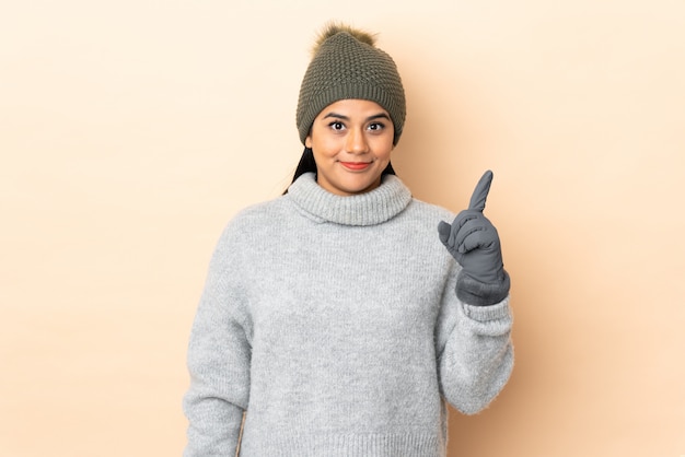 Giovane donna colombiana con cappello invernale isolato sul muro beige che punta con il dito indice una grande idea