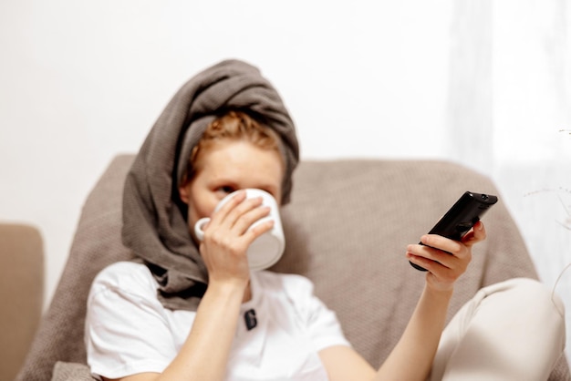 Giovane donna con abiti casual asciugamano sulla testa bevendo tè caldo e riposando guardando la TV Rilassarsi dopo aver fatto la doccia Tempo per prendersi cura di sé Rilassarsi a casa