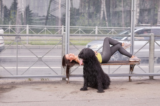 Giovane donna con briard nero vicino a lei è sdraiata su una panchina sulla stazione dei trasporti pubblici mentre aspetta l'autobus.