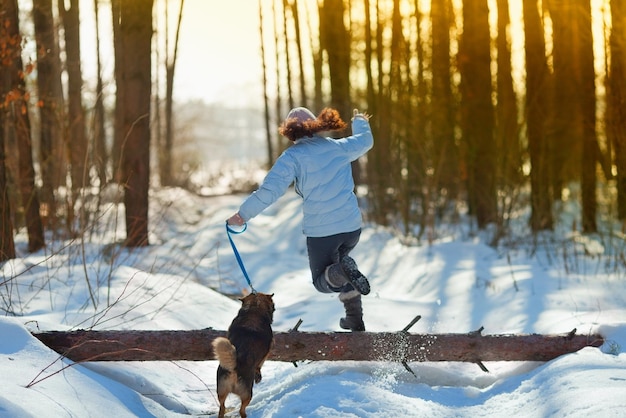 Giovane donna con cane che salta su un tronco nella foresta invernale innevata