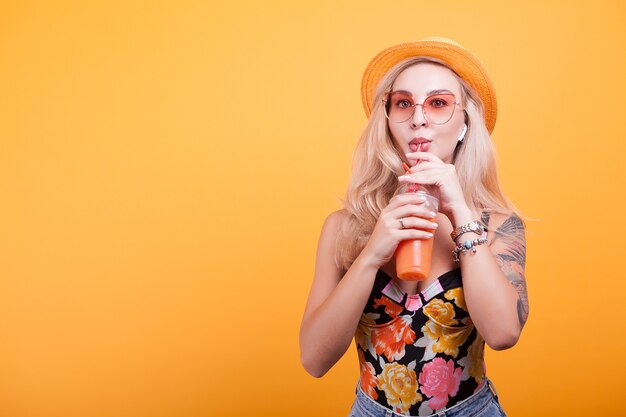 Giovane donna con capelli biondi che beve succo d'arancia con occhiali da sole in studio su sfondo giallo